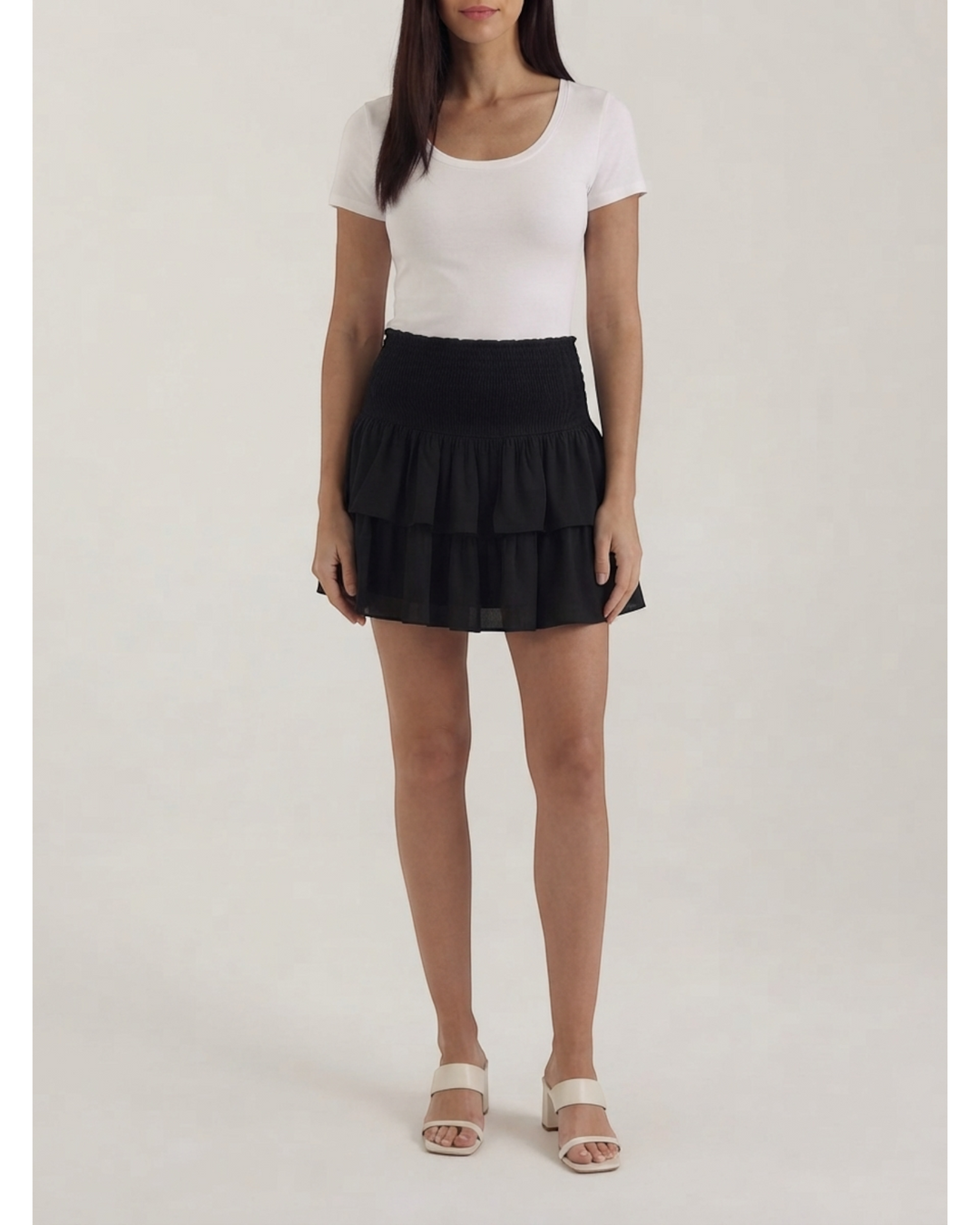 Luna Moda black mini skirt with a smocked high waist and layered ruffles worn by a woman with a white t-shirt