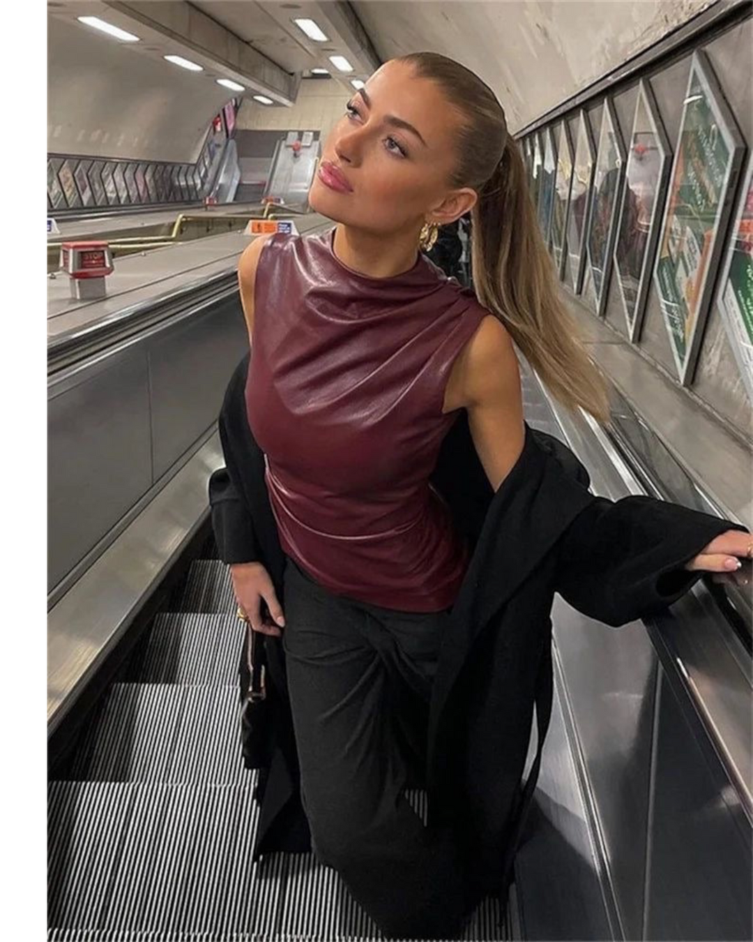 Luna Moda sleeveless burgundy faux leather top with a high neck, worn by a woman on an escalator in a city setting.