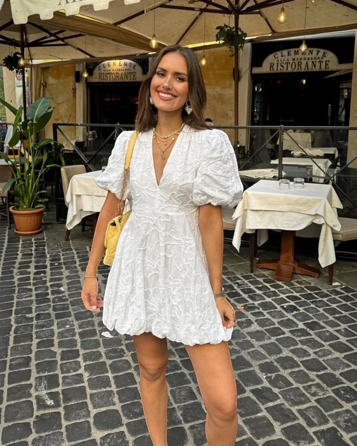 Luna Moda white mini dress with puff sleeves and delicate embroidery worn by a smiling woman at an outdoor cafe