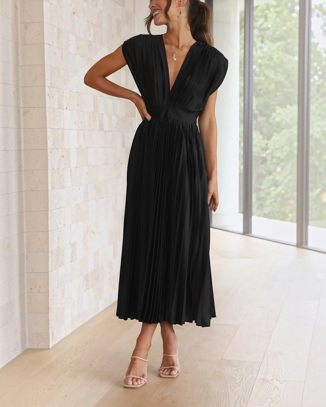 Luna Moda Isla black pleated midi dress with a deep V-neckline and high waist, worn by a woman in a bright room.