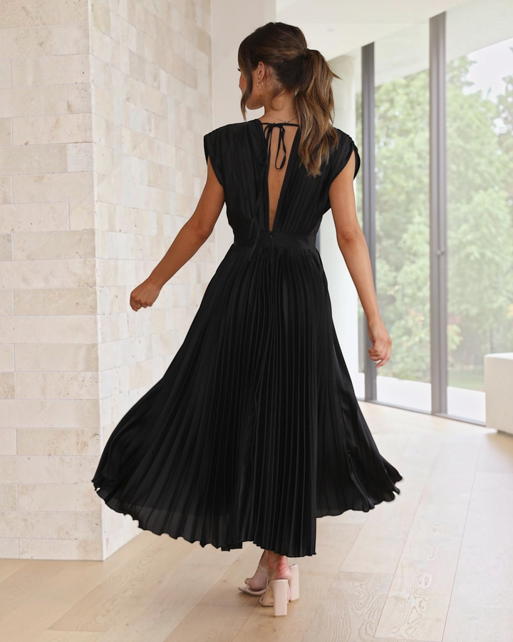 Luna Moda Isla black pleated midi dress with a deep V-back and tie detail, worn by a woman in a bright interior.