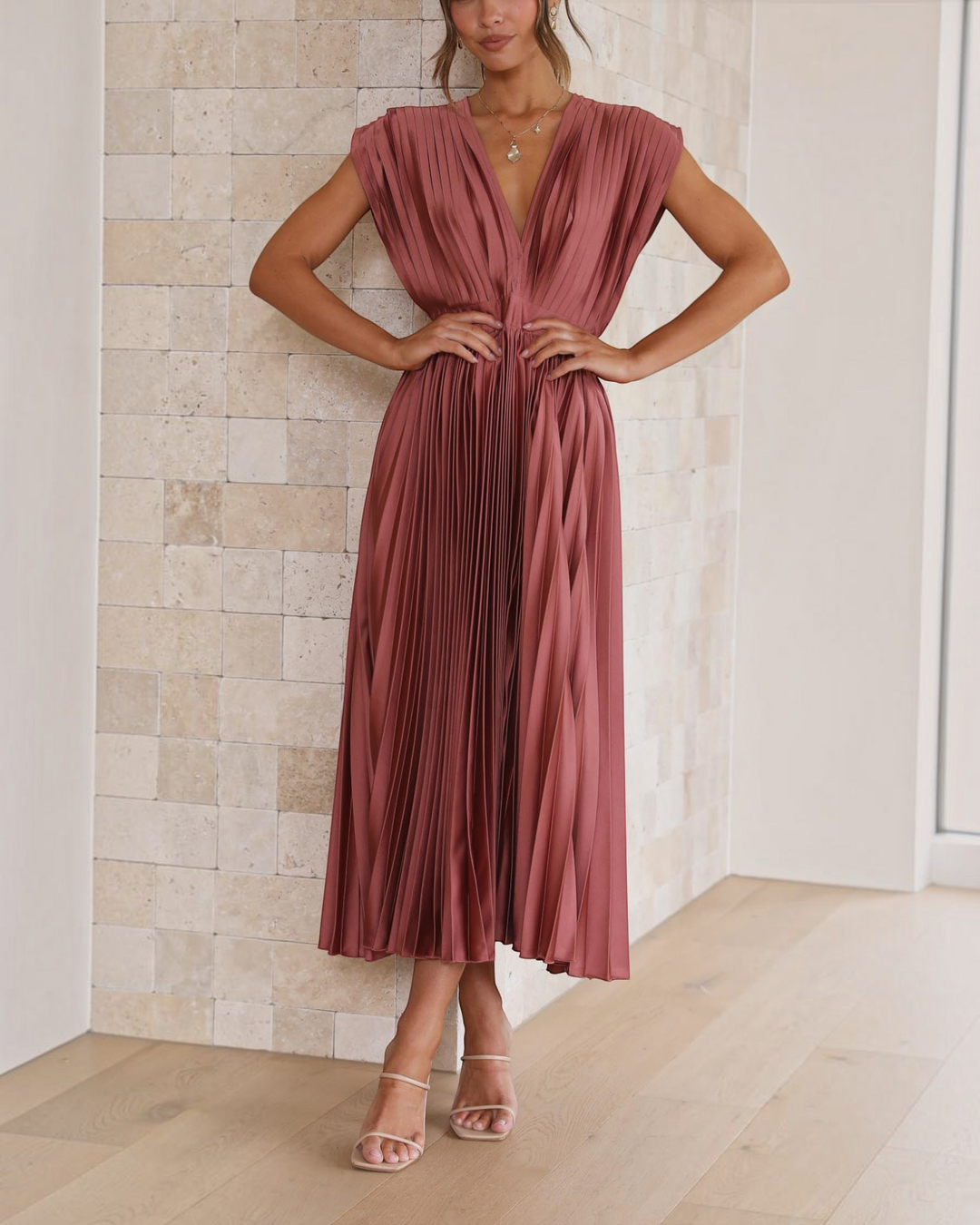 Luna Moda Isla rose red pleated midi dress with deep V-neck and high waist worn by a model against a stone wall.