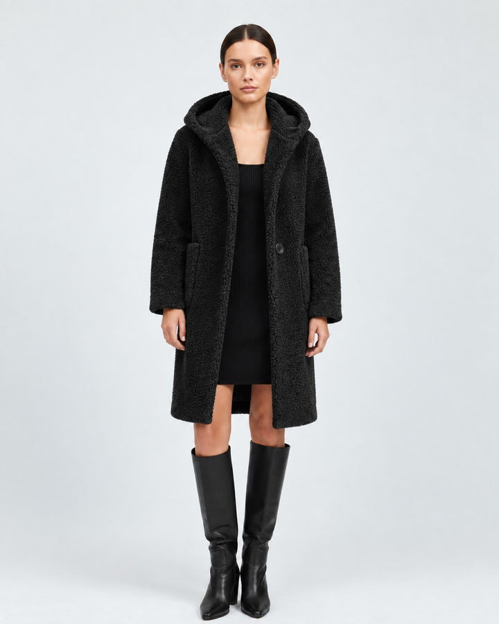 Model wearing a Luna Moda black knee-length hooded wool coat with patch pockets over a black dress and tall boots.