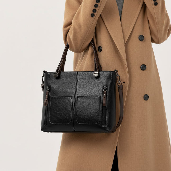 Luna Moda black leather handbag with dual front pockets and sturdy handles, worn by a woman in a camel coat.