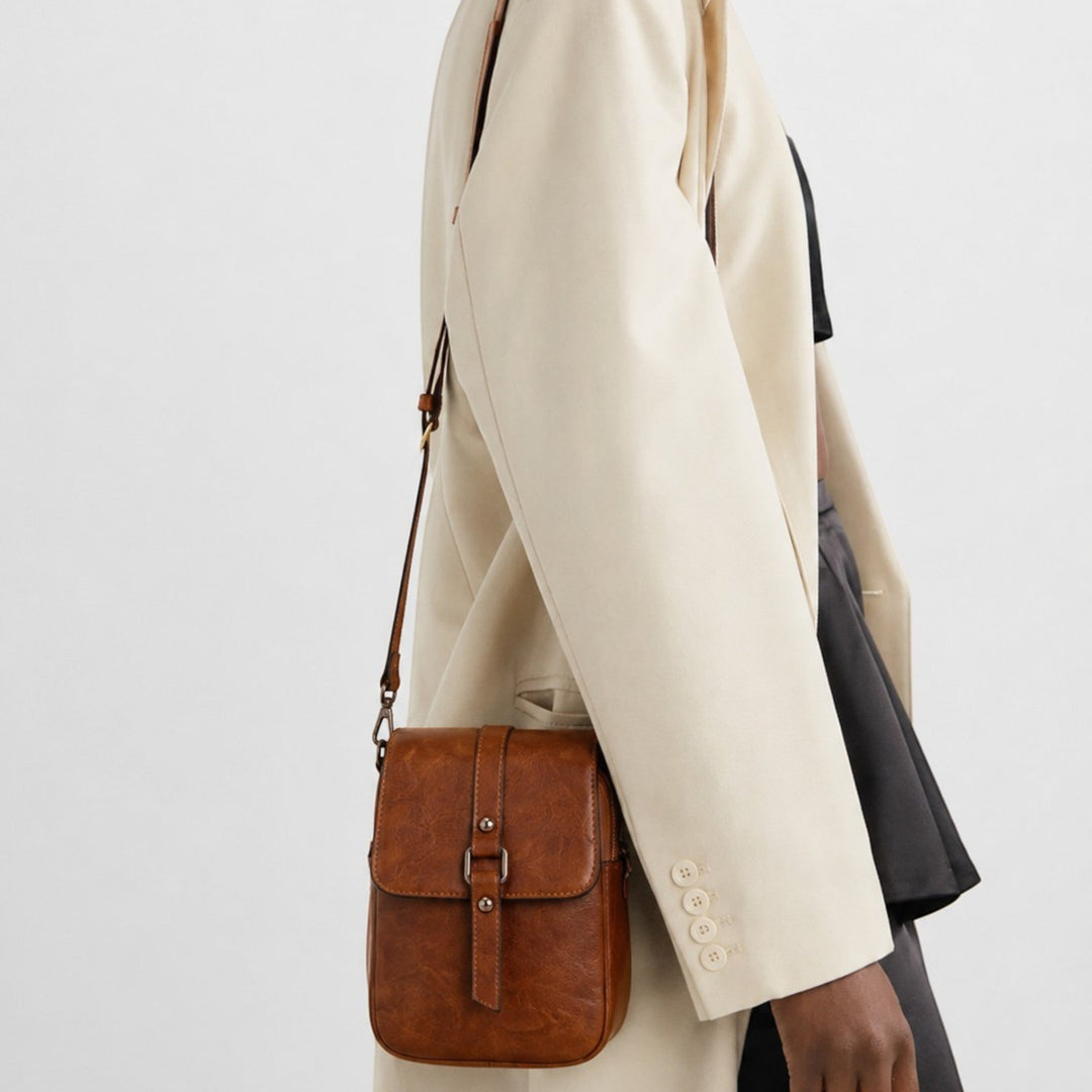 Luna Moda vertical crossbody bag in brown leather with a front buckle flap, worn by a model in a beige blazer.