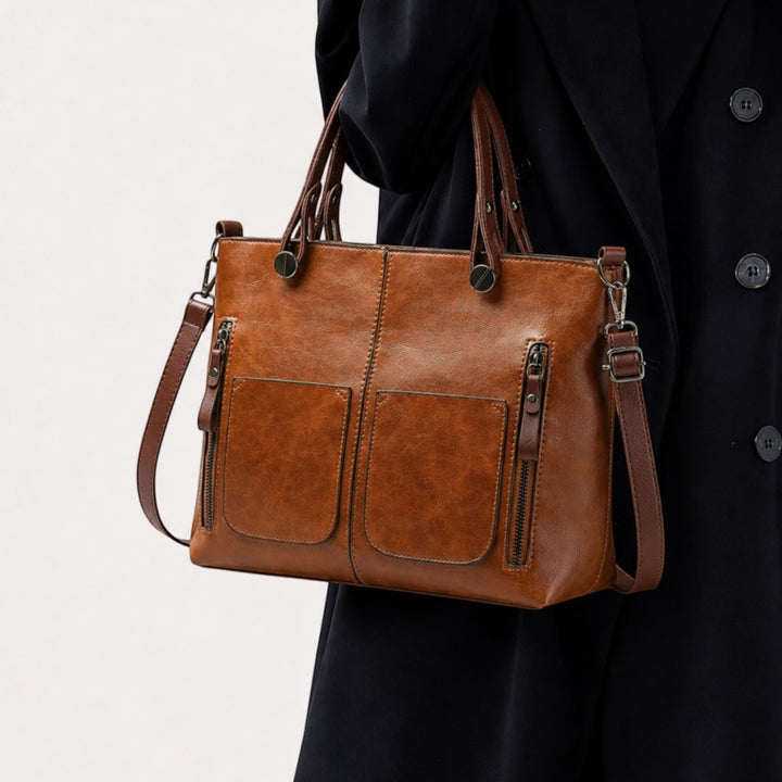 Luna Moda brown leather handbag with dual front pockets and sturdy handles, worn over a dark coat for a casual look.