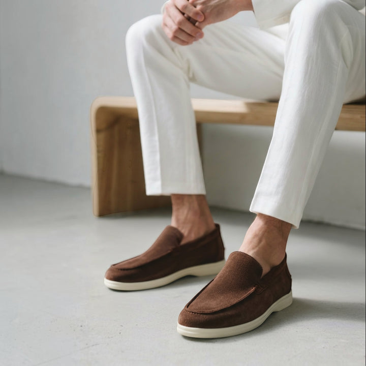 Luna Moda brown suede loafers with supportive white soles worn by a man in white trousers sitting on a wooden bench.