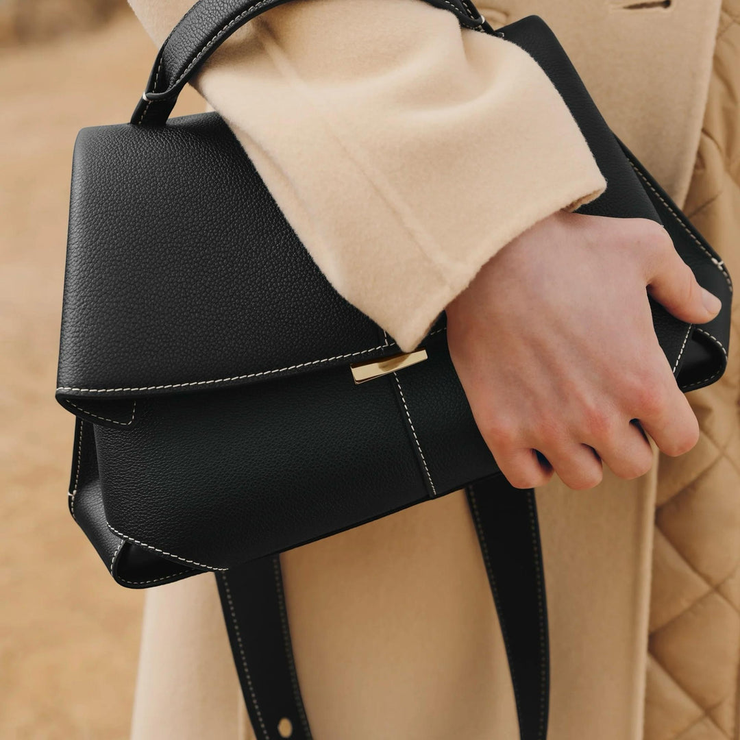 Luna Moda Lara black leather handbag with white contrast stitching and gold hardware held by a person in a beige coat.