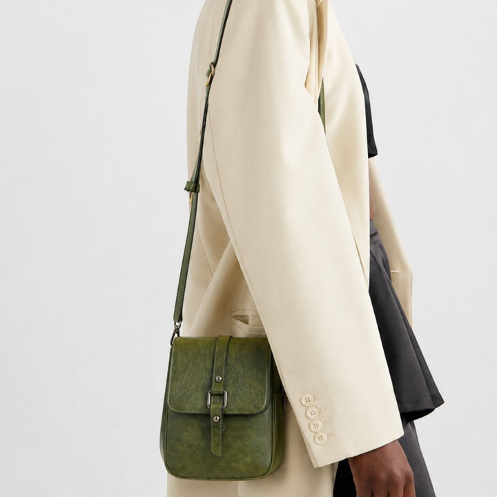 Luna Moda vertical crossbody bag in olive green with a front buckle flap, worn by a model in a cream blazer.