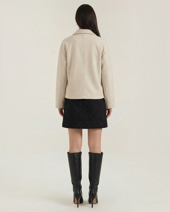 Back view of a woman wearing a Luna Moda beige zip-up jacket with a relaxed fit, paired with a black skirt and boots.